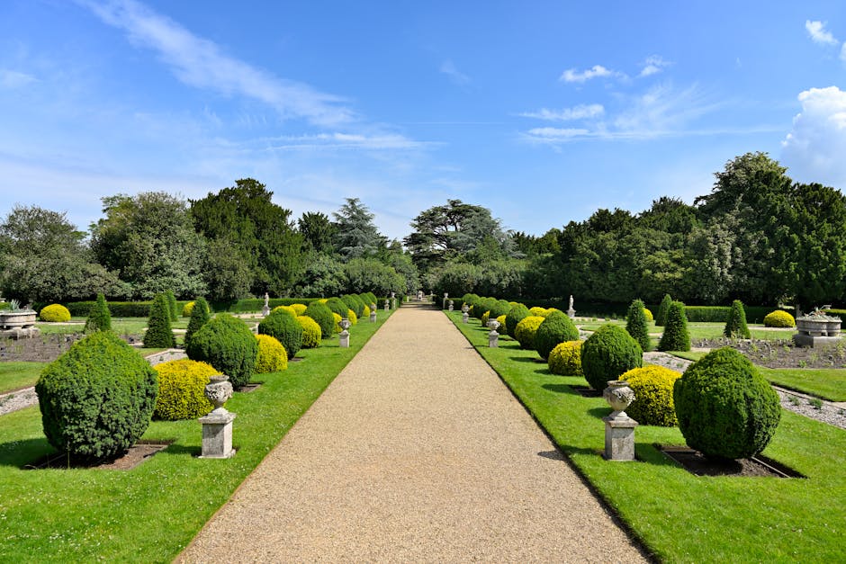 A symmetrical garden scene featuring a gravel pathway flanked by neatly trimmed, rounded boxwood bushes and small ornamental urns on stone pedestals. The pathway stretches into the distance, bordered 