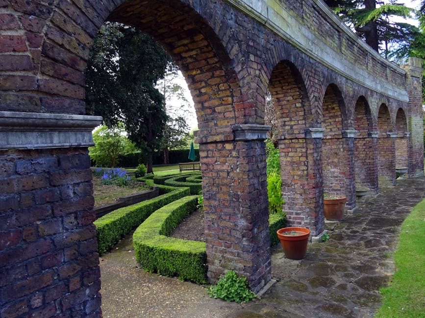 A historic brick archway with multiple arches runs along the length of a garden path, constructed from weathered reddish-brown bricks. Beneath the arches, a stone-paved walkway is bordered by neatly t