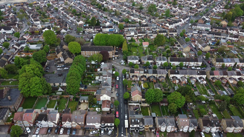 An aerial view of a residential suburban garden scene showing neatly maintained lawns, flowerbeds, and paved pathways. Several gardens feature lush green grass, some with trimmed hedges and small tree