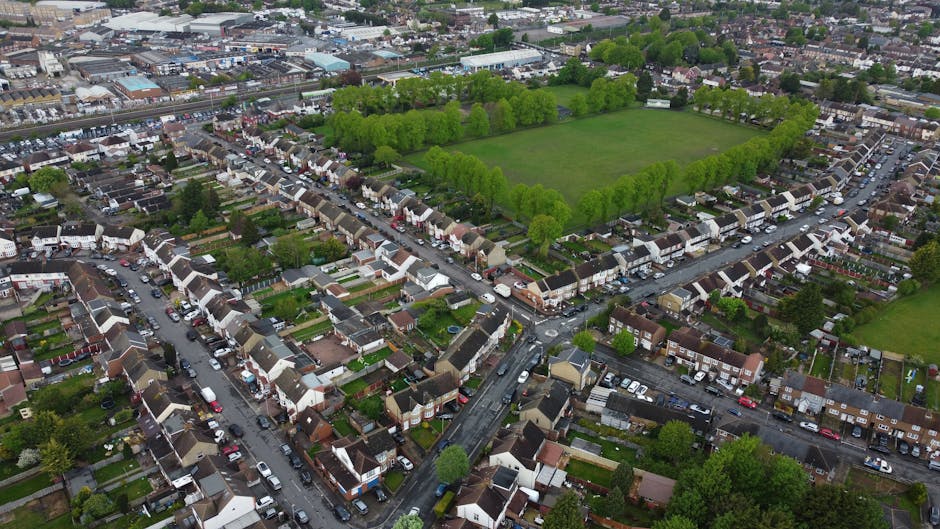 An aerial view of a residential suburban garden scene showing neatly maintained lawns, flowerbeds, and paved pathways. Several gardens feature lush green grass, some with trimmed hedges and small tree